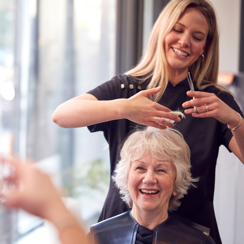 Eine ältere Dame bekommt die Haare von einer Frisöse geschnitten. Beide lachen und wirken sehr entspannt.