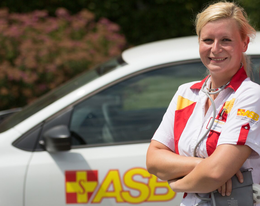 Zwei Mitarbeiter der ambulanten Pflege stehen vor ihrem Fahrzeug und lachen in die Kamera.