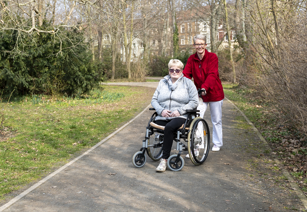 Eine Pflegerin schiebt eine Seniorin durch den Park. Beide lächeln freundlich in die Kamera. die Sonne scheint.