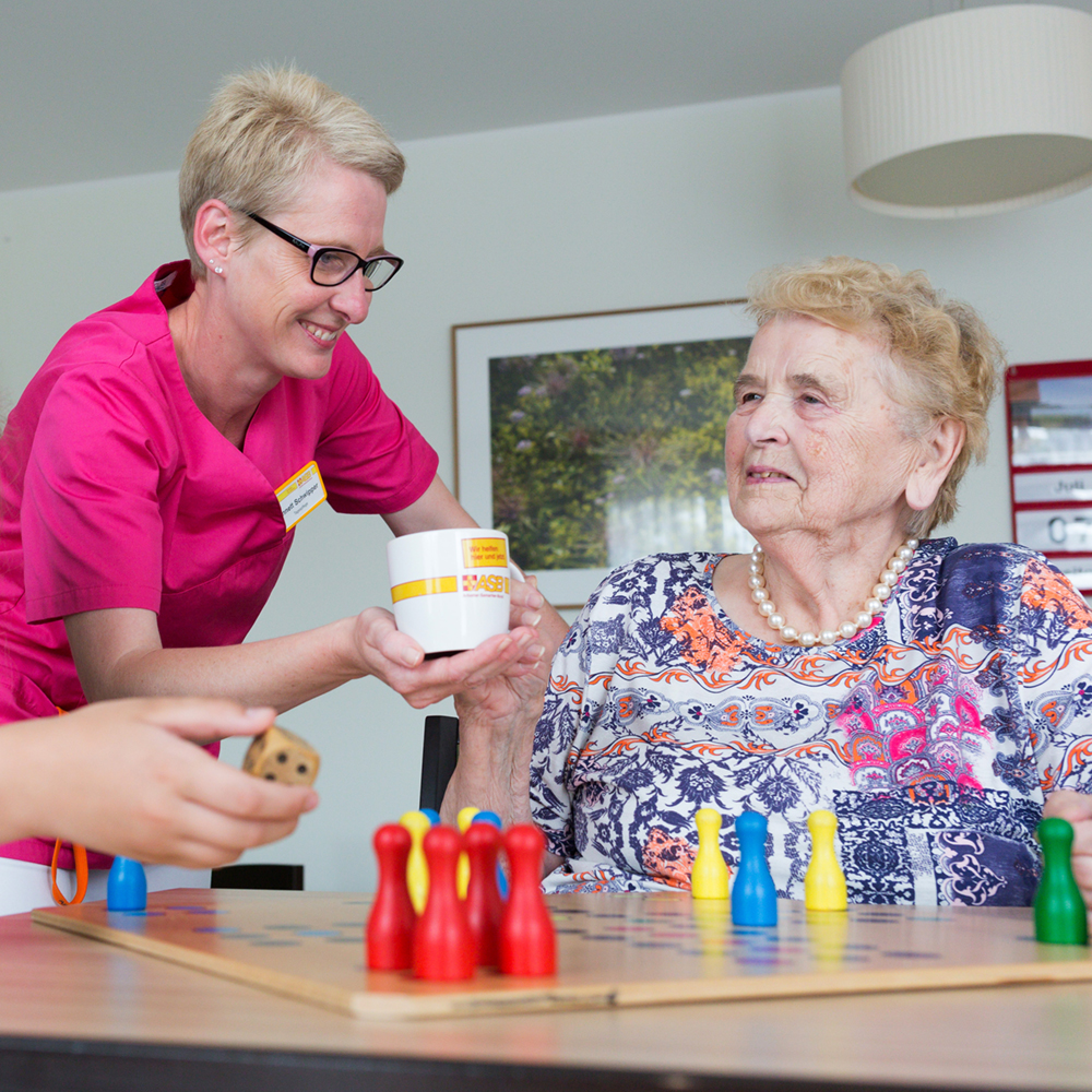 Eine ältere Dame bekommt von einer Pflegerin eine Tasse gereicht. Im Vordergrund befindet sich ein Tisch mit einem Spielbrett darauf.